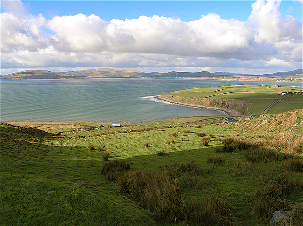 Ballinskelligs Bay on the west coast of Ireland with green fields in the foreground, mountains in the distance and clouds in a blue sky.