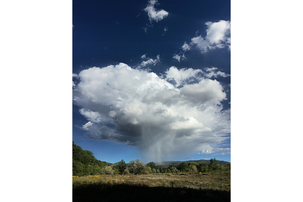 A sunny day in Taos with a massive rain cloud in a deep blue sky dropping rain that never touches the green landscape and mountains below.