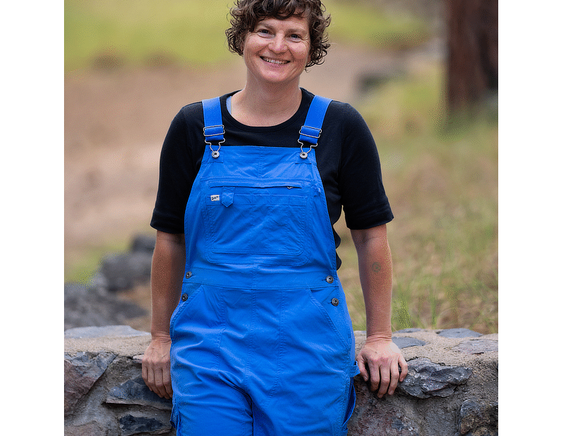 Image of a woman in blue bib overalls and black shirt leaning on a low stone wall with grass in the background.