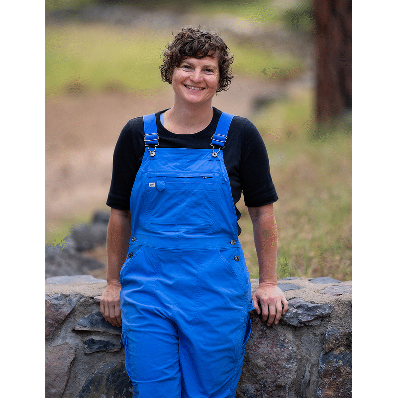 Image of a woman in blue bib overalls and black shirt leaning on a low stone wall with grass in the background.