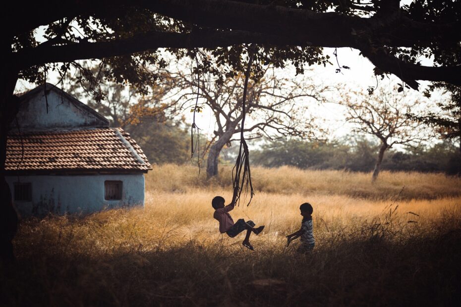 silhouette of two children on a tree swing with grass and a building in the background