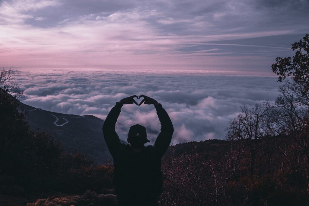 Silhouette of a person on a mountain holding their hands overhead in the shape of a heart against a distant background filled with clouds.