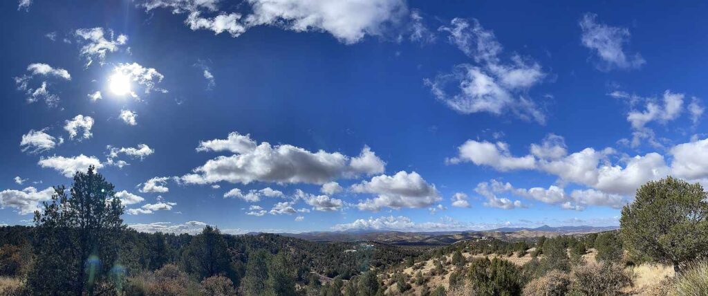 Panoramic image of bright blue skies with puffy white clouds and a bright sun above a landscape with trees and mountains in southwestern New Mexico near Silver City.