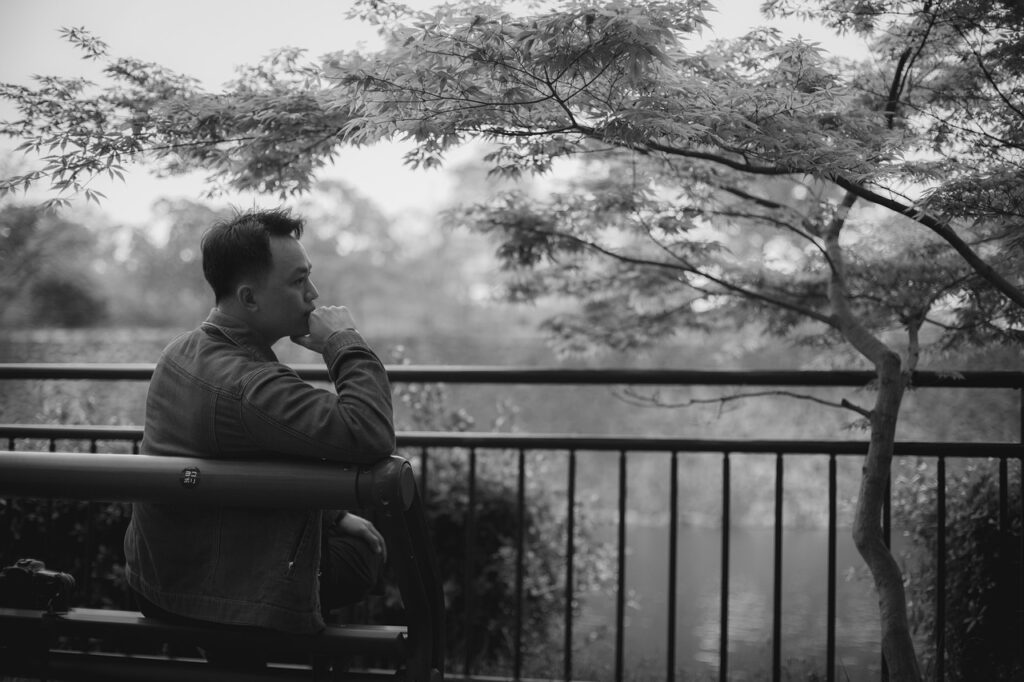 Black and white image of a man sitting on a park bench looking to the right toward tree branches with a river in the background.